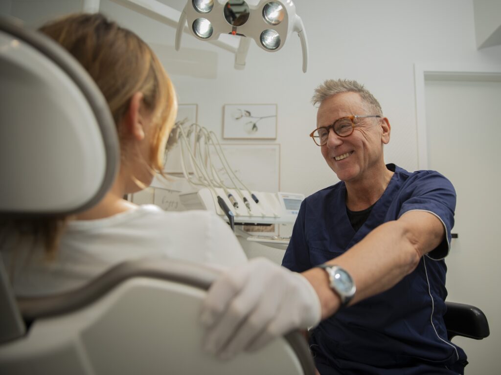 Ein älterer Zahnarzt mit Brille und Handschuhen lächelt freundlich zu einer Patientin