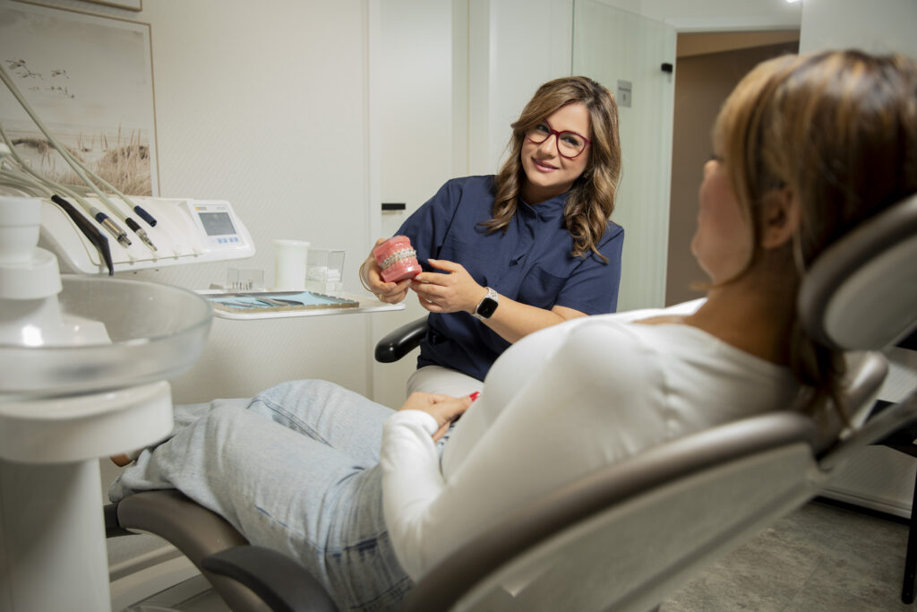 Eine Frau mit Brille und blauer Bluse zeigt einer Patientin freundlich ein Zahnmodell