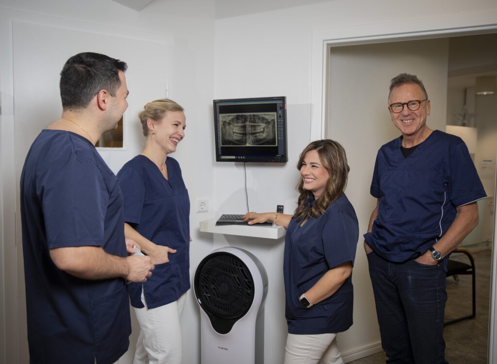 Vier freundlich lachende Zahnärztinnen und Zahnärzte in blauer Kleidung vor Röntgenbild